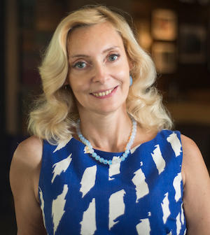 Headshot of Professor Karina Korostelina. She is smiling at the camera wearing a blue and white dress and beaded necklace.