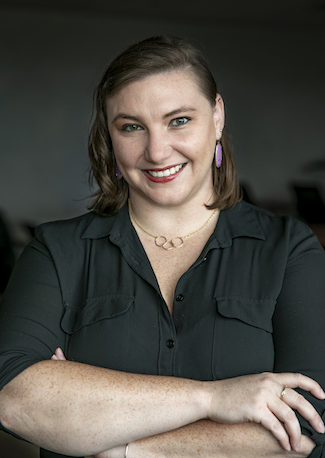 PhD Candidate Emily Sample stands with her arms crossed and smiles at the camera. She is wearing a black collared shirt and a golden necklace with three interconnected circles.