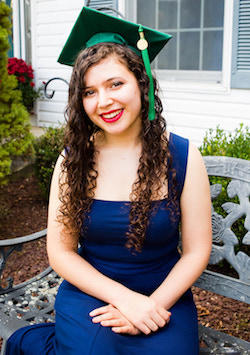 Photo of Carter School graduate Lily Krietzberg sitting on a stone bench in a blue dress with a green Mason Mortar Board cap on her head.
