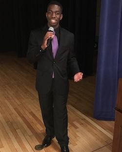 Picture of Carter School bachelor's graduate Phillip Robinson, a young Black man, dressed in a suit and holding a microphone in a stage setting.