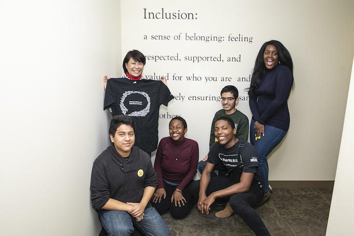 Students pose in front of a mural with a definition of inclusion: "a sense of belonging: felling respected, supported, and valued for who you are and (unreadable) ensuring the same for others."