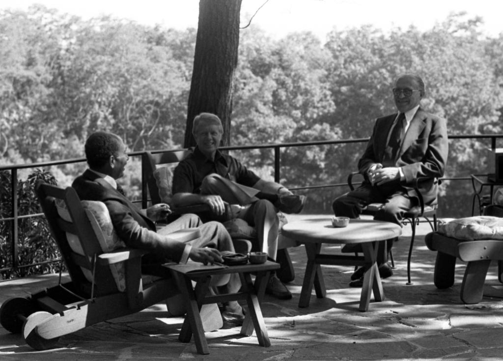 Black and white photo of Anwar Sadat, Jimmy Carter, and Menachem Begin chatting on a balcony overlook at Camp David