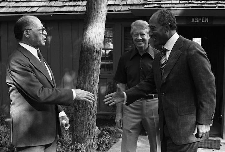 Black and white photo of Menachem Begin and Anwar Sadat going to shake hands upon arrival at Camp David. Jimmy Carter smiles in the background.