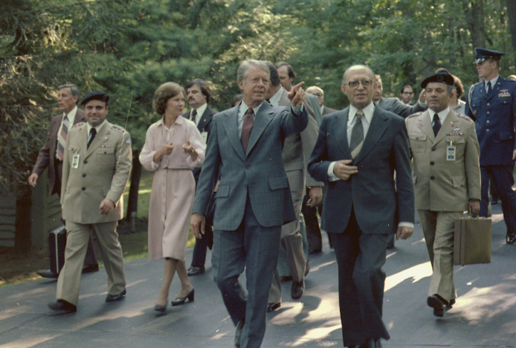 Photo of Jimmy Carter and Menachem Begin walking at Camp David. Rosalynn Carter is in the background talking to someone from Begin's entourage. Jimmy Carter is pointing something out to Begin.