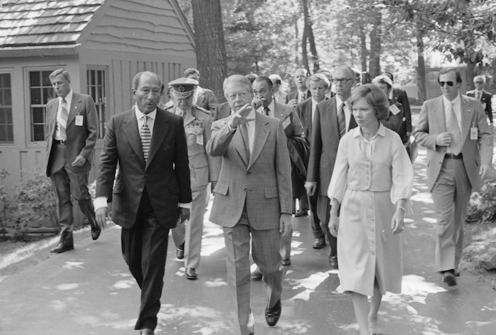 Black and white photo of Anwar Sadat, Jimmy Carter, and Rosalynn Carter walking at Camp David with their entourages. Carter is pointing out something to Sadat as they walk.