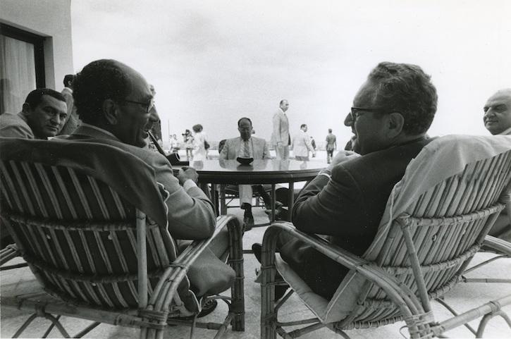 Black and white photo of Egyptian President Anwar Sadat and Henry Kissinger chatting at the head of a round table. At the other end in the distance is Harold Saunders.