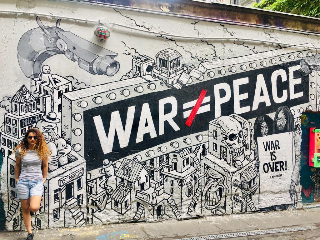 Fatma Jabbari, a Carter School PhD student, stands in front of an intricate street mural in Berlin that features a large block of text reading "WAR [DOES NOT EQUAL] PEACE"