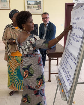Two women wearing traditional Congolese clothes stand and write on a large piece of poster paper as they analyze and vote on next steps in the peace program.
